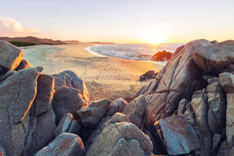 Felsen im Vordergrund, Sandstrand und Meer bei Sonnenaufgang im Hintergrund
